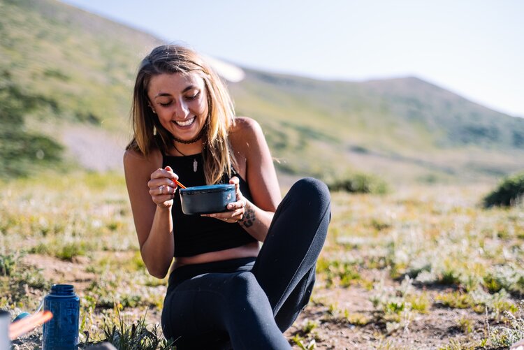 Woman sitting in the ground in the nature and smiling while eating food from the small pot